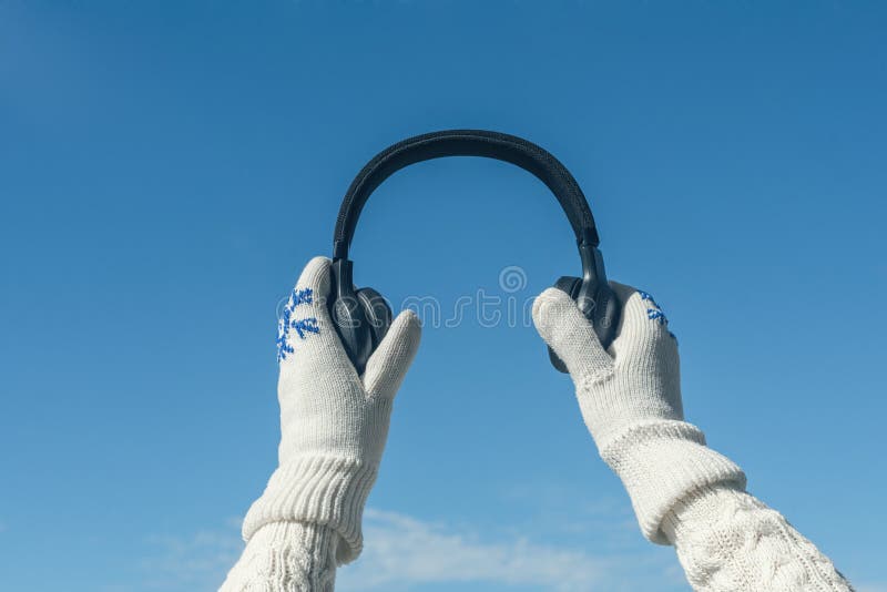 Female hands in the winter mittens with a headphones on the blue sky background