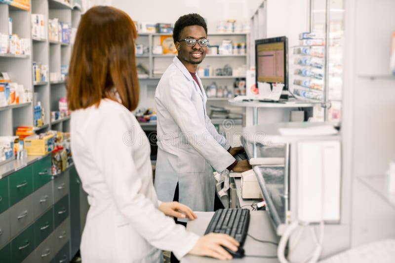 Female and male pharmacists in modern pharmacy, looking at each other, while working on computers.