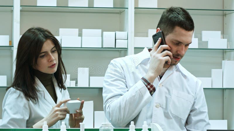 Female and male pharmacists working in pharmacy, talking via smart phone and checking pills royalty free stock photo Female and male pharmacists working in pharmacy, talking via smart phone and checking pills royalty free stock photo