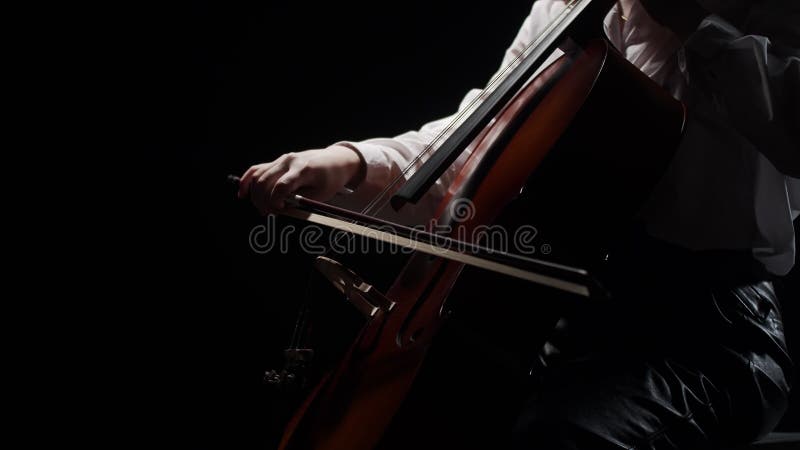 Female musician plays classical music on cello while sitting on chair in studio, dark background, side view. Woman stock video