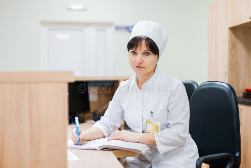 A female nurse in a white medical gown sits at a wooden desk at the reception desk and makes entries in a work log. Woman doctor