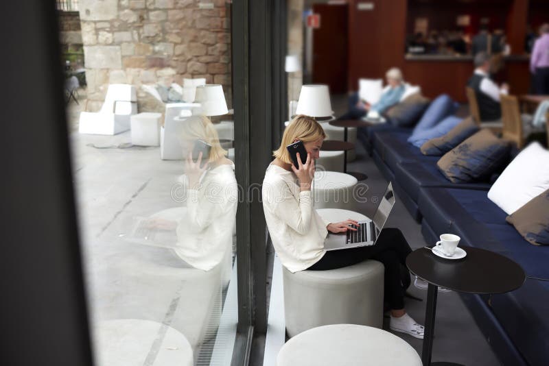 Young businesswoman sitting front open laptop computer and talk on smartphone, female person using net-book during morning breakfast in modern coffee shop, freelancer working on the distance in cafe