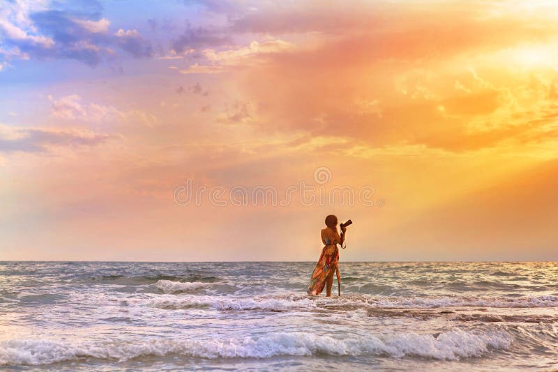 Female photographer with professional camera standing in the sea and takes picture of sunset. Female in the dress in the waves royalty free stock photos Female photographer with professional camera standing in the sea and takes picture of sunset. Female in the dress in the waves royalty free stock photos