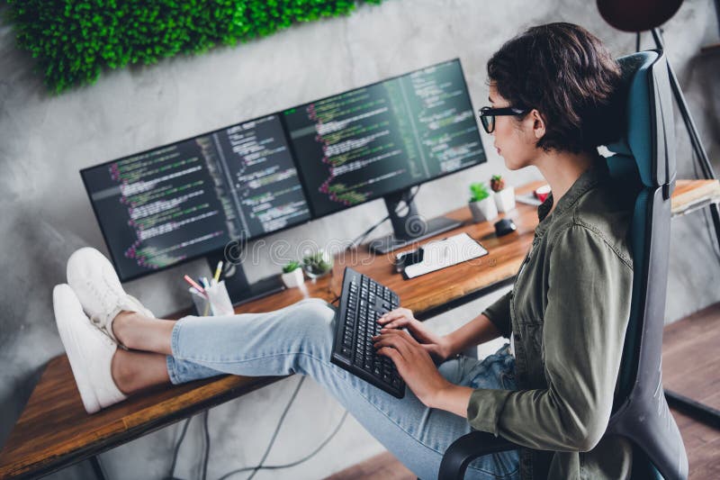 Female programmer in casual style focusing on computer screens while working from her home office in a modern workspace stock photo