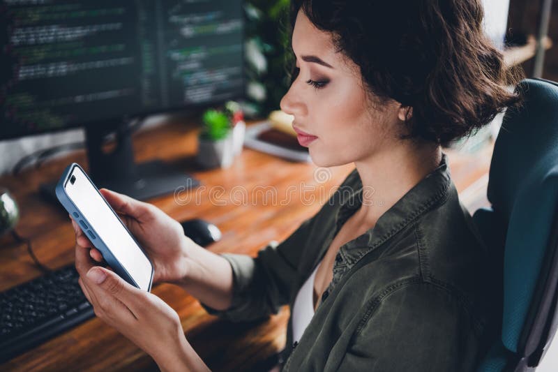Female programmer using smartphone while working at her desk, coding on a computer in a stylish home workspace stock image