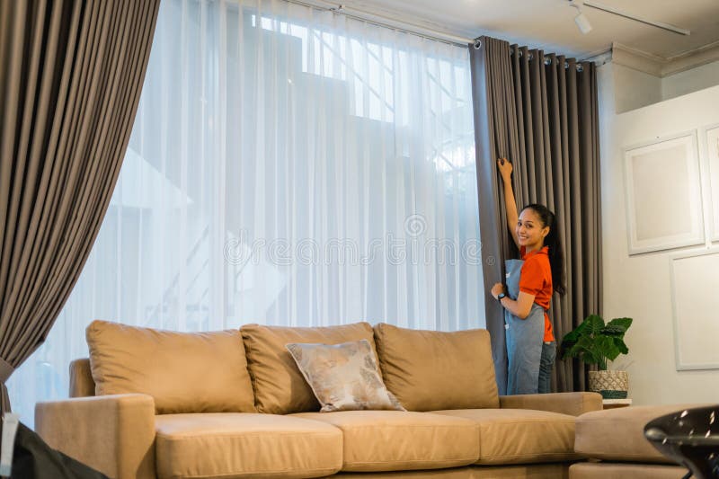 Female shop assistant in apron opening the window curtain behind a sofa. In a furniture store