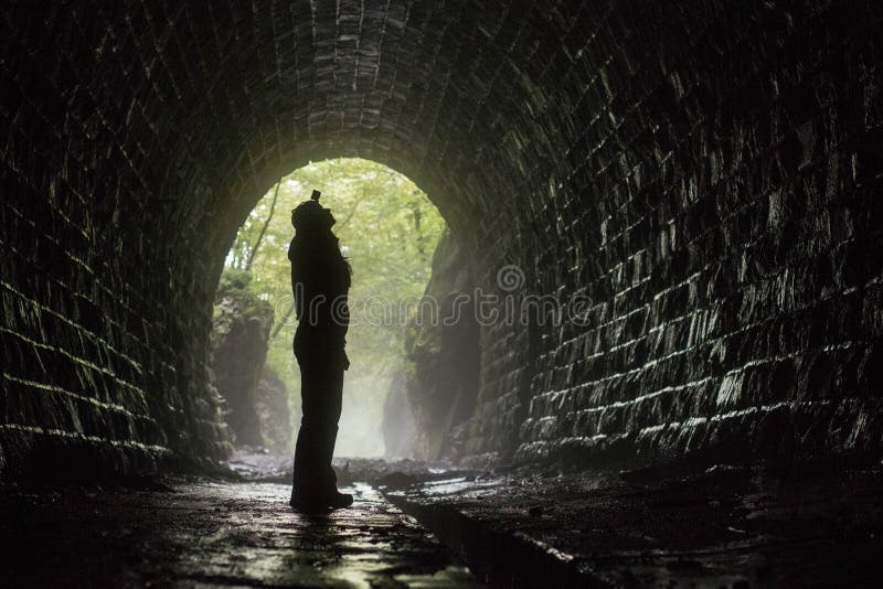 Female silhouette with headlamp in wet brick tunnel, green forest background