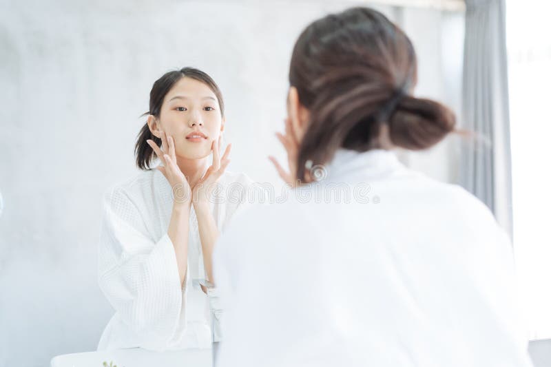 Female Skin Care. Young asian woman touching her face and looking to mirror in bathroom.