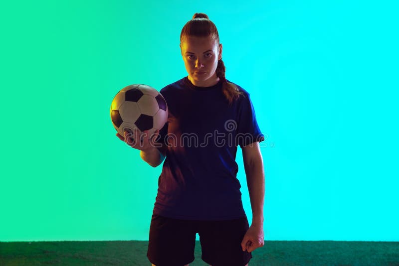 Young female soccer or football player with long hair in sportwear holding ball, posing confident on gradient background, neon. Concept of healthy lifestyle, professional sport, motion, movement. Young female soccer or football player with long hair in sportwear holding ball, posing confident on gradient background, neon. Concept of healthy lifestyle, professional sport, motion, movement.