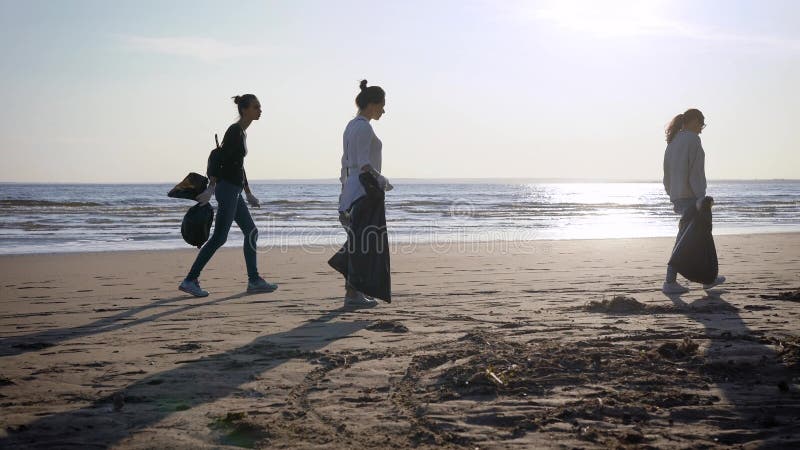 Female volunteers are walking with plastic bags over wild sand beach in daytime, cleaning city territory