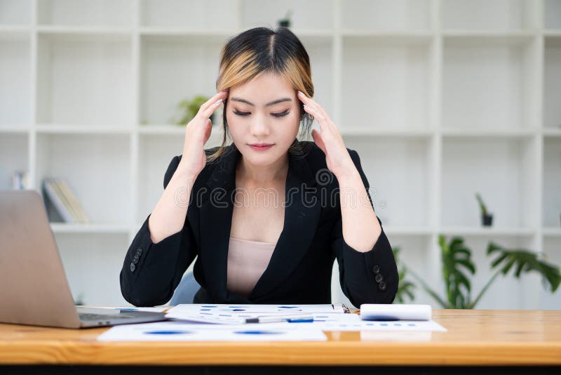 Femeie asiatică cu dureri de cap care lucrează la un laptop și analizează hărți financiare pe masă royalty free stock image