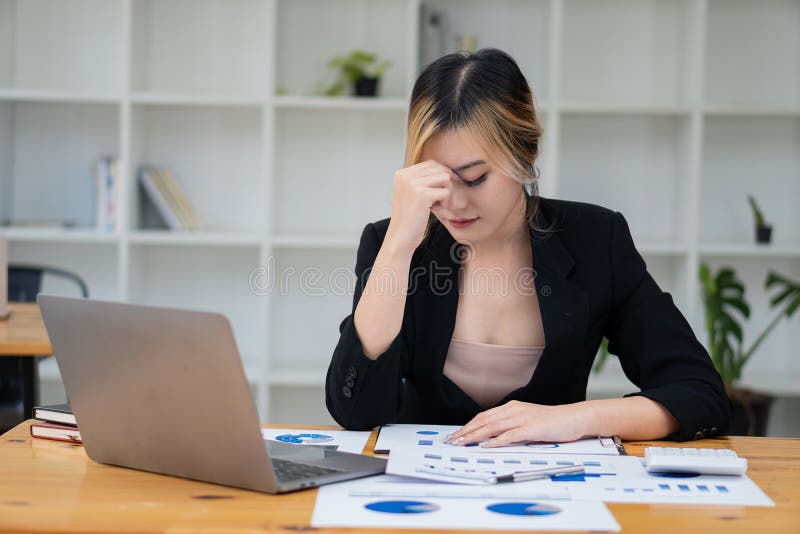 Femeie asiatică cu dureri de cap care lucrează la un laptop și analizează hărți financiare pe masă stock photography