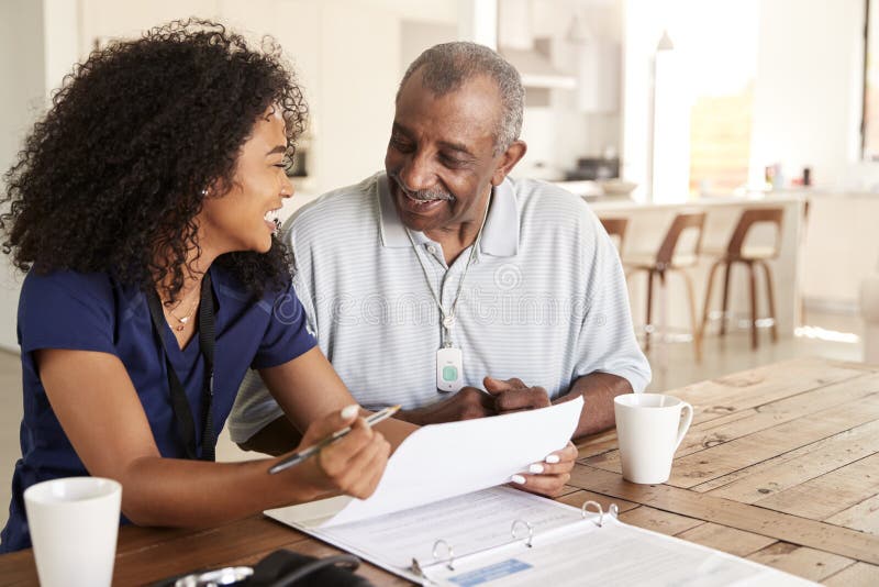 Femeie care se ocupă cu asistența medicală fericită și care stă la masă și zâmbește cu un bărbat în vârstă în timpul unei vizite l stock image