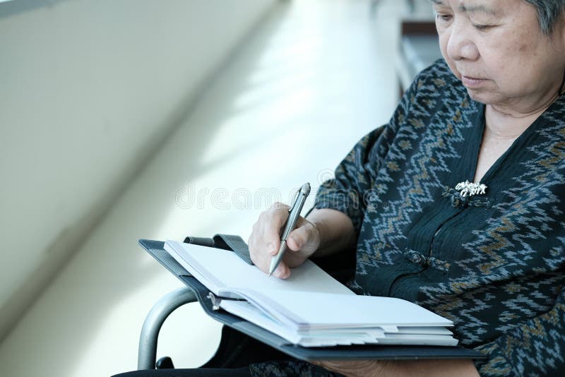Femeie mai în vârstă pe scaun cu rotile nota de scris pe notebook-ul de lângă fereastră. royalty free stock images