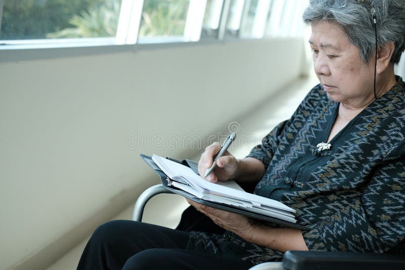 Femeie mai în vârstă pe scaun cu rotile nota de scris pe notebook-ul de lângă fereastră. royalty free stock photos