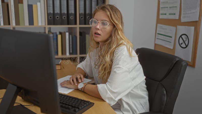 Femeie tânără care lucrează într-un birou cu un computer înconjurat de documente și rafturi cu aspect concentrat și profesional royalty free stock photo
