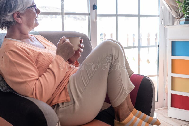 Femeie în vârstă relaxată care stă acasă pe fotoliu și bea o ceașcă de ceai. doamna cu părul gri care se uită pe fereastră royalty free stock image