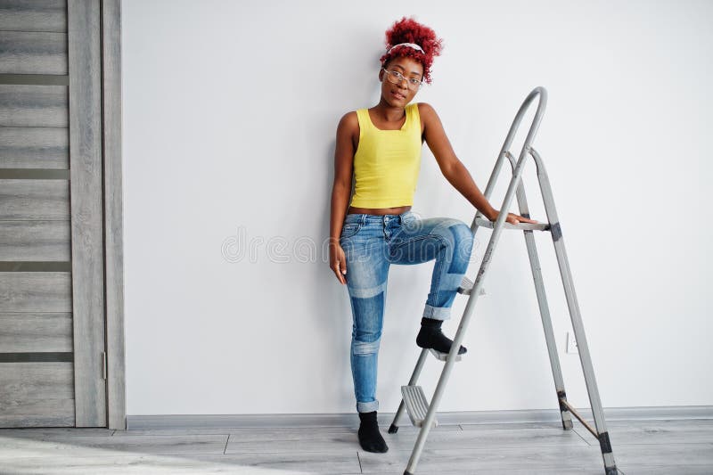 Femme afro-américaine en singlet jaune et lunettes contre mur blanc stand om échelle en fer photographie stock