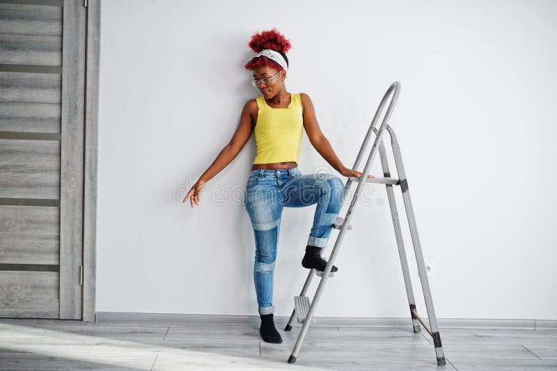 Femme afro-américaine en singlet jaune et lunettes contre mur blanc stand om échelle en fer photographie stock libre de droits