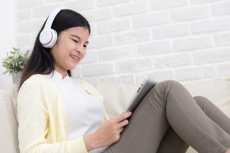 Femme asiatique de sourire à l'aide de la tablette et écoutant la musique sur le sofa à la maison dans le salon images libres de droits Femme asiatique de sourire à l'aide de la tablette et écoutant la musique sur le sofa à la maison dans le salon images libres de droits