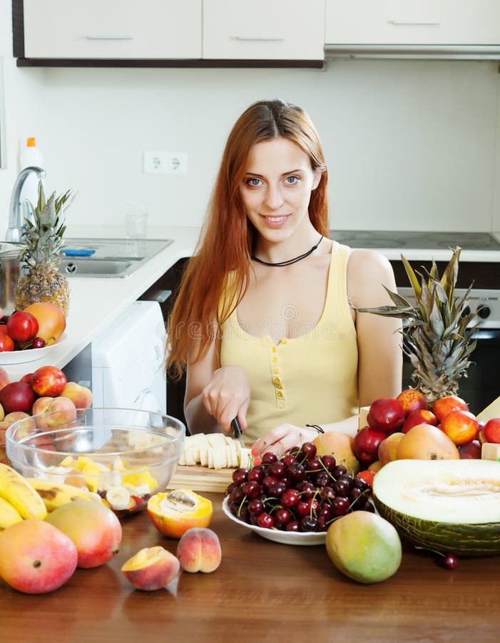 Femme aux cheveux longs positive faisant la salade de fruits coupée images stock Femme aux cheveux longs positive faisant la salade de fruits coupée images stock