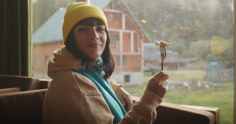 Femme avec un morceau de nourriture sur une fourchette souriant par une fenêtre avec vue. ambiance intérieure chaleureuse avec lum banque de vidéos