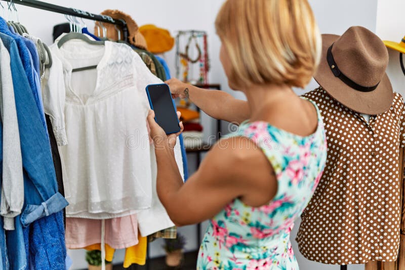 Femme blonde d'âge moyen faire une photo par le smartphone à vêtements dans la boutique de vêtements