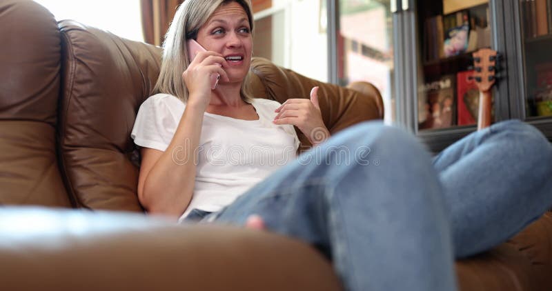 Femme blonde mature se détend sur canapé en cuir parlant sur smartphone banque de vidéos