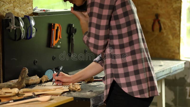 Femme charpentier joyeuse discutant des détails de commande avec son client sur smartphone et prend des notes banque de vidéos