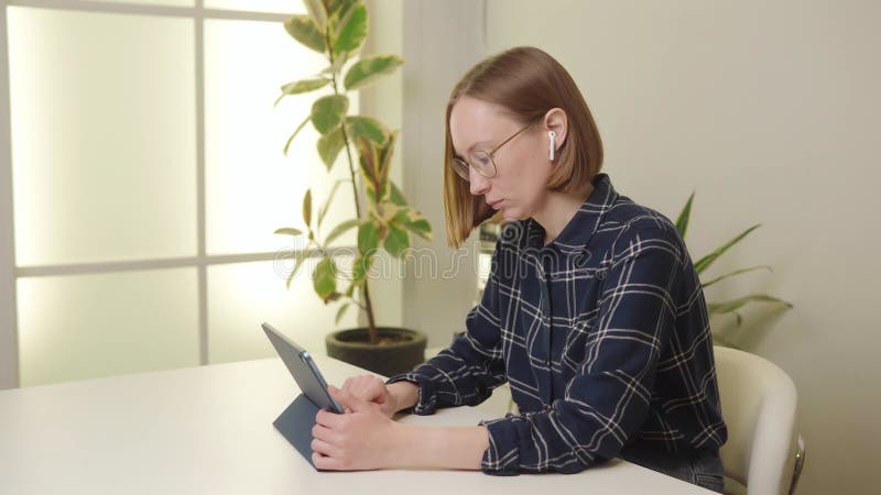 Femme ciblée en chemise à carreaux à l'aide d'une tablette sur un bureau blanc avec une plante en pot en arrière-plan. banque de vidéos
