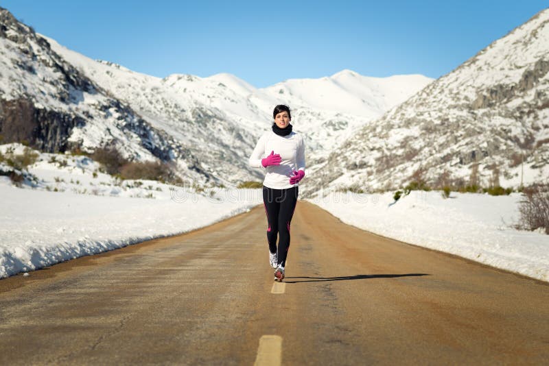 Female athlete running in cold winter mountain road. Woman training for marathon outdoors wearing warm sportswear. Female athlete running in cold winter mountain road. Woman training for marathon outdoors wearing warm sportswear.