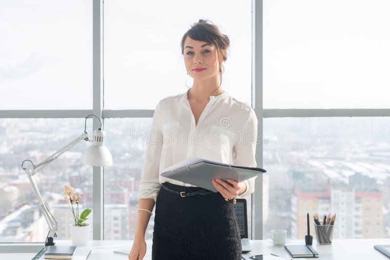 Femme d'affaires ambitieuse attirante se tenant dans le bureau moderne, tenant le dossier de papier, regardant l'appareil-photo, photographie stock Femme d'affaires ambitieuse attirante se tenant dans le bureau moderne, tenant le dossier de papier, regardant l'appareil-photo, photographie stock