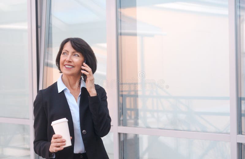 Femme d'affaires m? re ayant une coupure devant un immeuble de bureaux, parlant au t? l? phone et buvant du caf? photo libre de droits