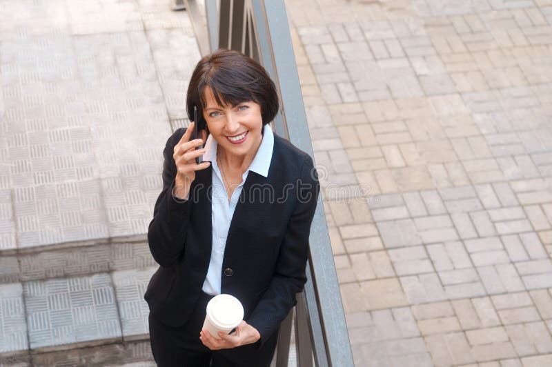 Femme d'affaires mûre ayant une coupure devant un immeuble de bureaux, parlant au téléphone et buvant du café photographie stock libre de droits