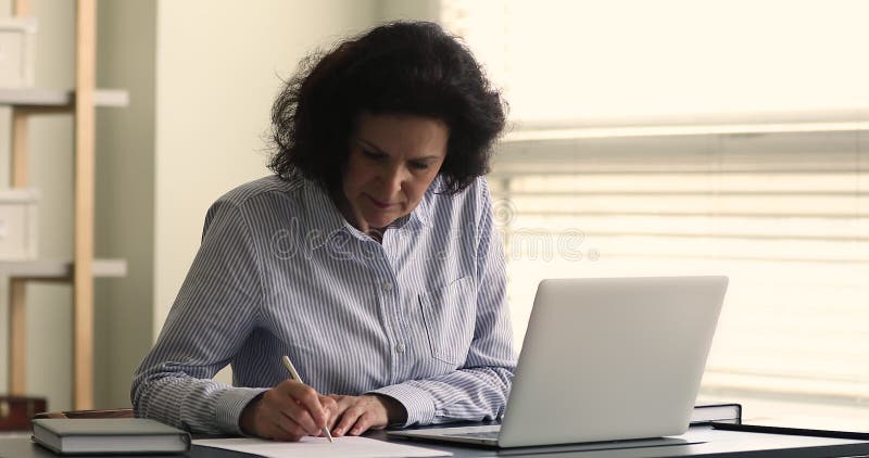 Femme d'affaires âgée assise au bureau textos utiliser ordinateur portable prend des notes banque de vidéos