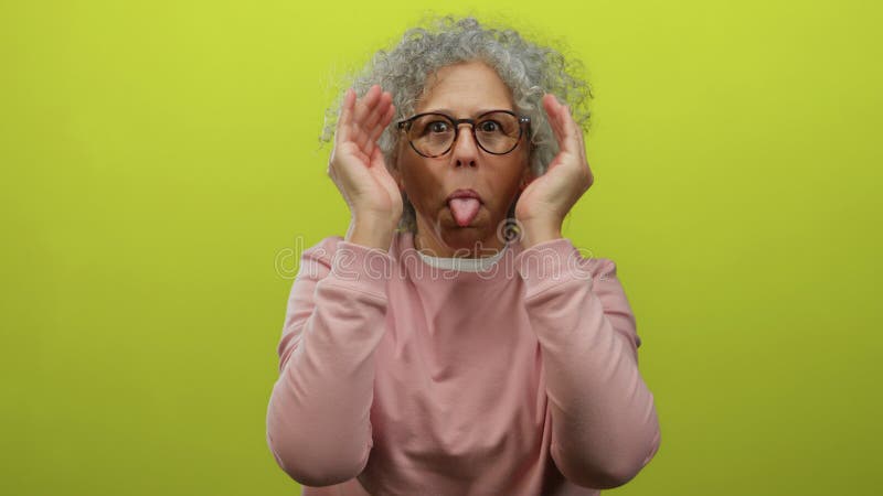 Femme d'âge moyen avec cheveux gris en pull rose enlève les mains du visage souriant devant fond jaune vif photos stock