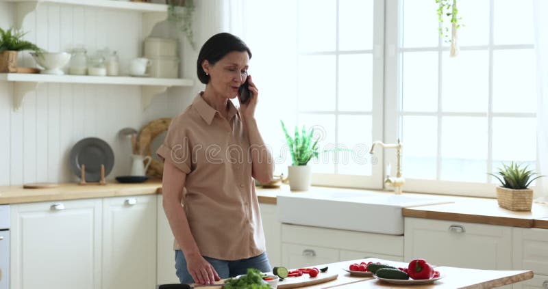 Femme d'âge moyen heureuse parlant sur téléphone portable en cuisine banque de vidéos