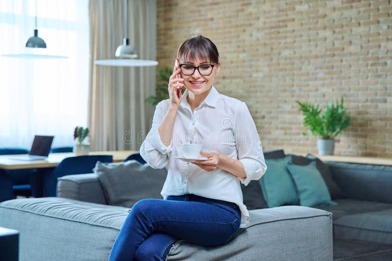 Middle aged woman talking on phone, having cup of coffee at home on couch in living room. Mature female resting, talking with friends, colleagues, family, leisure communication concept. Middle aged woman talking on phone, having cup of coffee at home on couch in living room. Mature female resting, talking with friends, colleagues, family, leisure communication concept