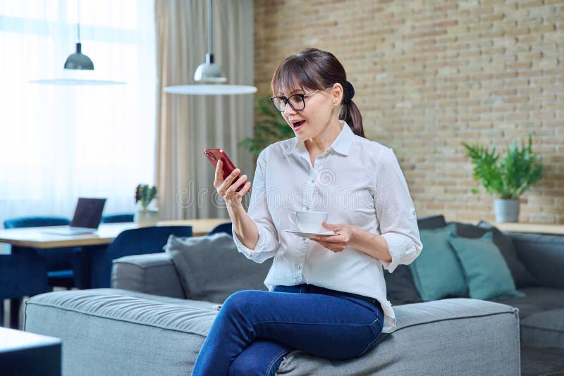 Middle aged woman using smartphone, with cup of coffee at home on couch. Mature female resting reading news, social networks, chatting with friends, colleagues, family, surprise joy happiness emotions. Middle aged woman using smartphone, with cup of coffee at home on couch. Mature female resting reading news, social networks, chatting with friends, colleagues, family, surprise joy happiness emotions