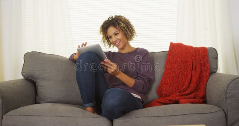 Une femme noire heureuse est assise sur un canapé en utilisant une tablette photo libre de droits