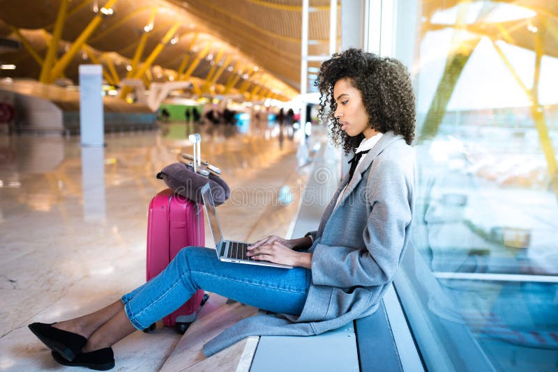 Femme de couleur travaillant avec l'ordinateur portable à l'aéroport attendant aux WI photographie stock Femme de couleur travaillant avec l'ordinateur portable à l'aéroport attendant aux WI photographie stock