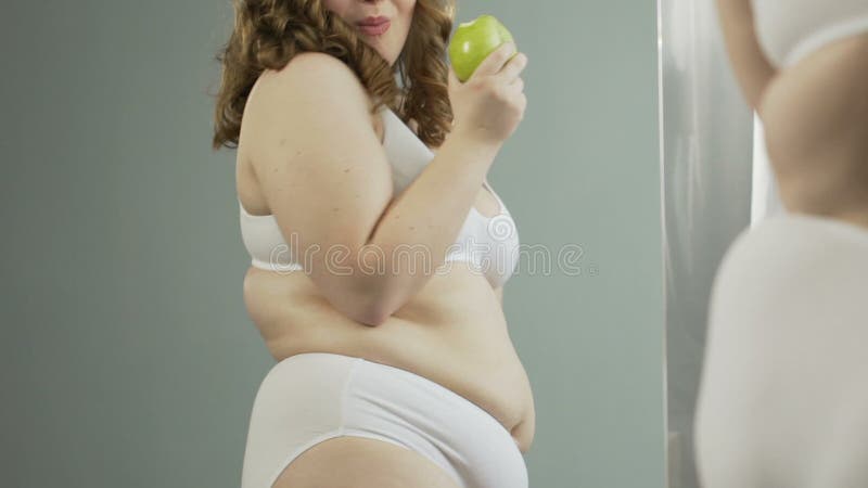 Femme de poids excessif dans les sous-vêtements admirant son corps dans le miroir et mangeant la pomme verte banque de vidéos