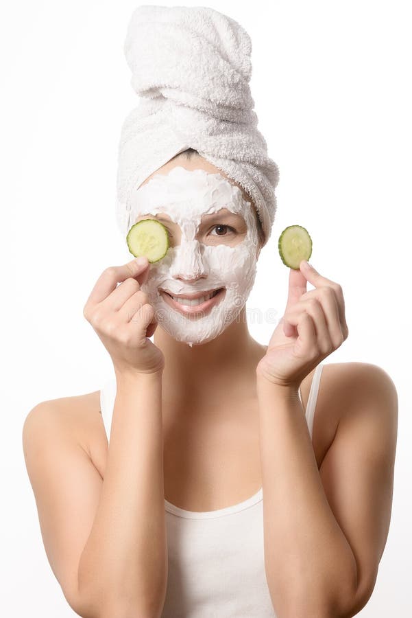 Smiling woman with her hair tied up in a white towel and a deep cleansing nourishing face mask applied to her skin holding a refreshing slice of cucumber to her eye in a beauty and skincare concept. Smiling woman with her hair tied up in a white towel and a deep cleansing nourishing face mask applied to her skin holding a refreshing slice of cucumber to her eye in a beauty and skincare concept