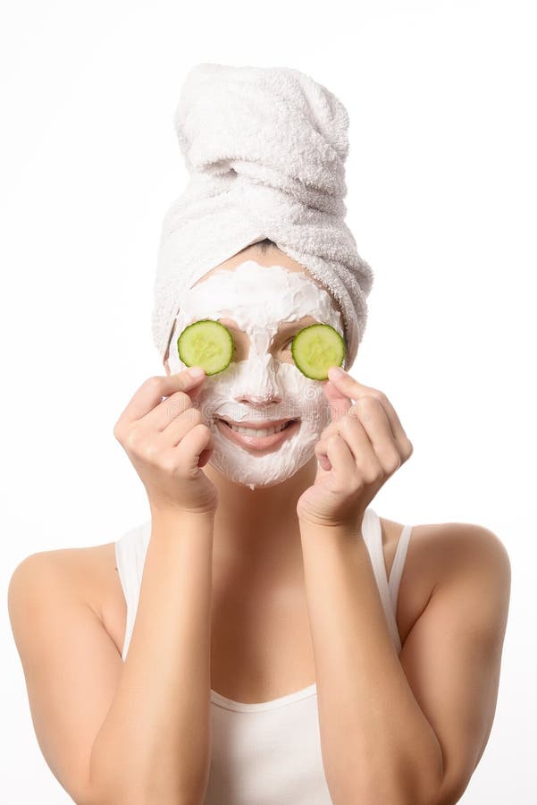 Smiling woman with her hair tied up in a white towel and a deep cleansing nourishing face mask applied to her skin holding a refreshing slice of cucumber to her eye in a beauty and skincare concept. Smiling woman with her hair tied up in a white towel and a deep cleansing nourishing face mask applied to her skin holding a refreshing slice of cucumber to her eye in a beauty and skincare concept