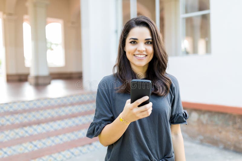 Femme de sourire tenant Smartphone tout en se tenant contre la construction image stock Femme de sourire tenant Smartphone tout en se tenant contre la construction image stock