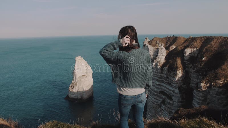 Femme de touristes enthousiaste heureuse rétroviseur à regarder la vue sur le rocher d'Étretat, touchant les cheveux soufflant au clips vidéos