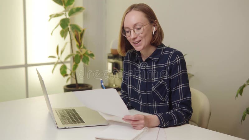 Femme donnant une conférence en regardant dans la caméra avec un ordinateur portable et des documents sur un bureau blanc. clips vidéos