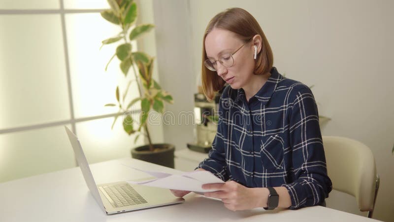 Femme en chemise bleue lunettes oreilles bourgeons examiner la paperasse avec un ordinateur portable ouvert à côté d'elle dans une clips vidéos
