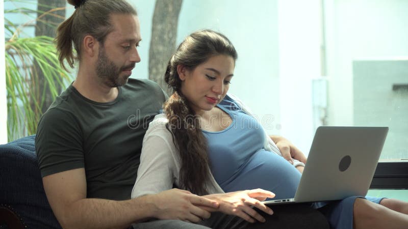 Femme enceinte assise sur un canapé avec mari utilisant un ordinateur portable ensemble dans le salon à la maison. concept couple clips vidéos