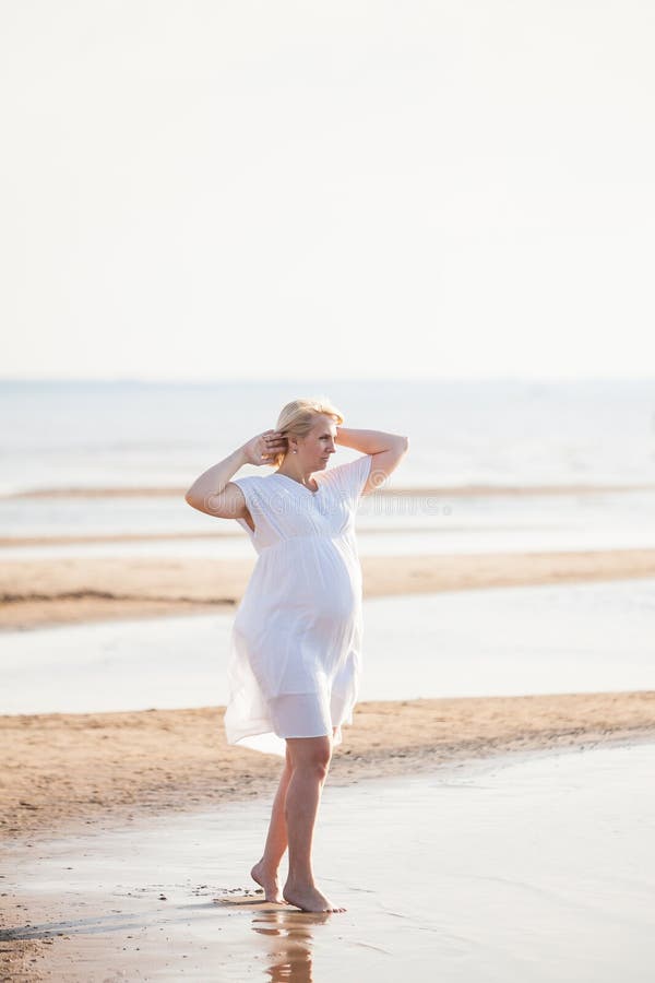 Femme enceinte dans la robe blanche posant sur la plage à la soirée ensoleillée Haute verticale principale images stock Femme enceinte dans la robe blanche posant sur la plage à la soirée ensoleillée Haute verticale principale images stock